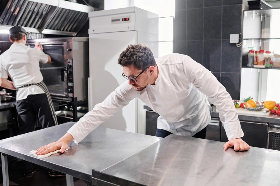 A chef in chef whites cleaning a stainless steel commercial bench with a microfibre cloth